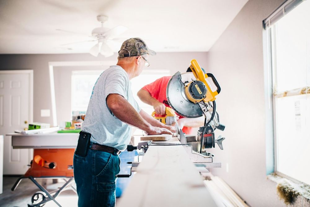 A handyman cutting a board at a miter saw inside a bright room under renovation